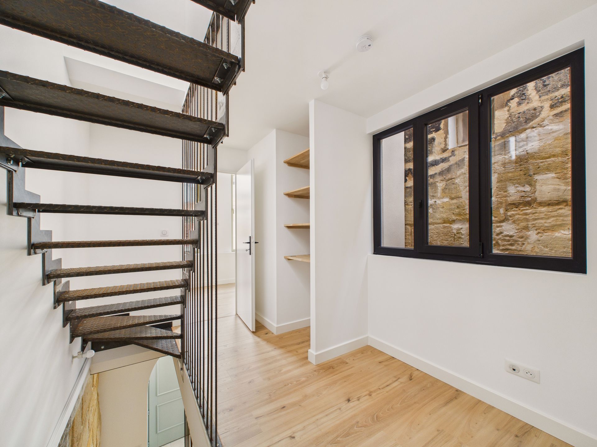 Vue sous un escalier noir, à côté de fenêtres donnant sur un mur en pierre.
