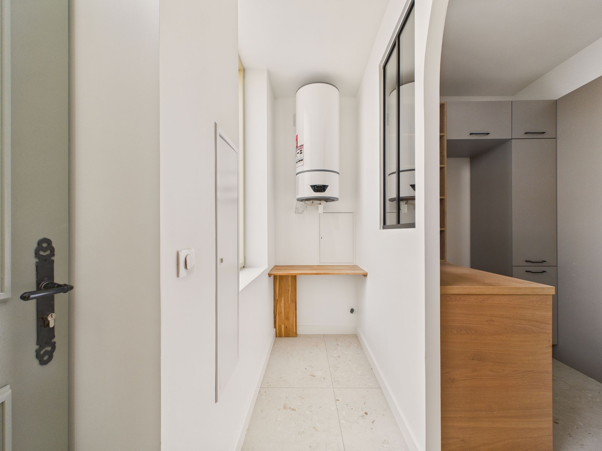 Couloir blanc ouvrant sur une cuisine, avec un chauffe-eau dans une niche.