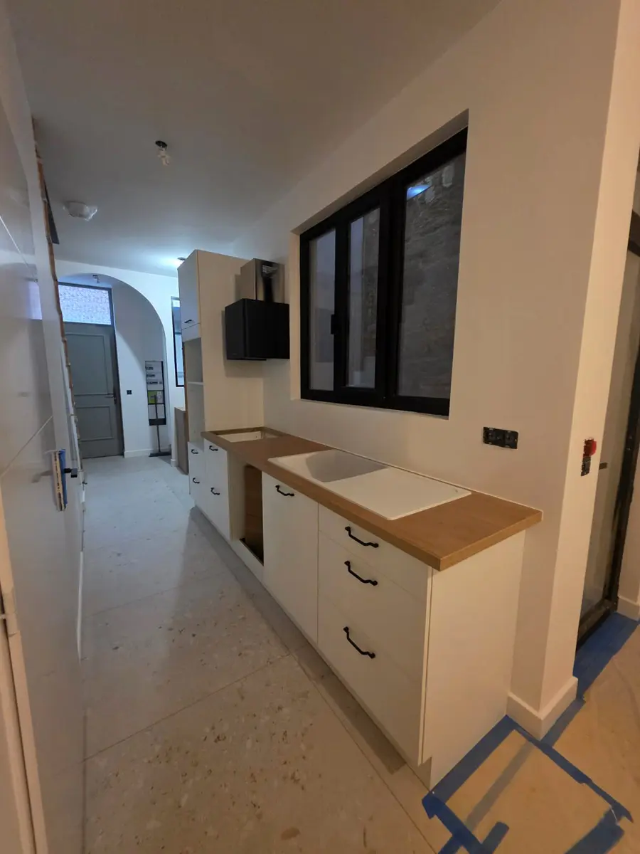 Cuisine blanche à plan de travail en bois, séparée par une verrière noire.