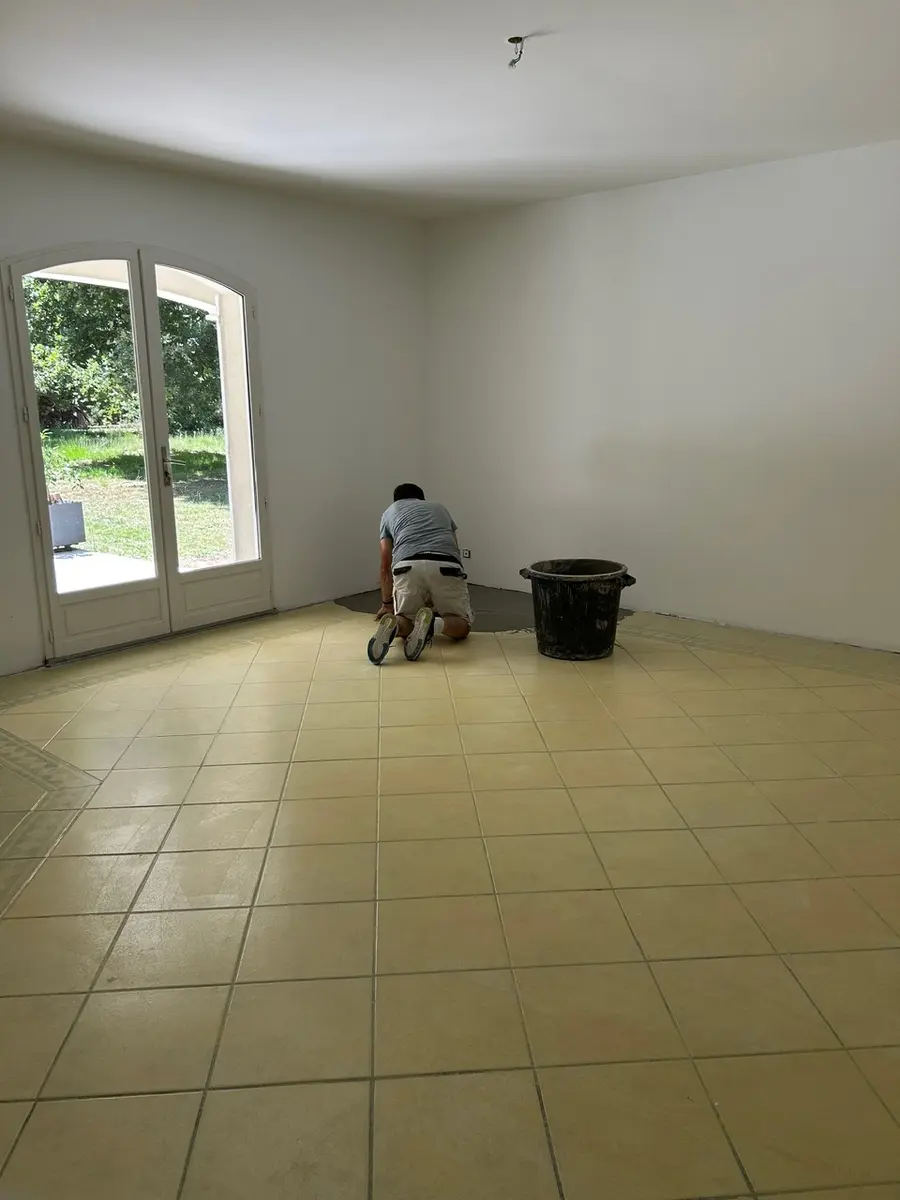 Grande pièce en travaux avec un sol carrelé et du matériel près d’une fenêtre.