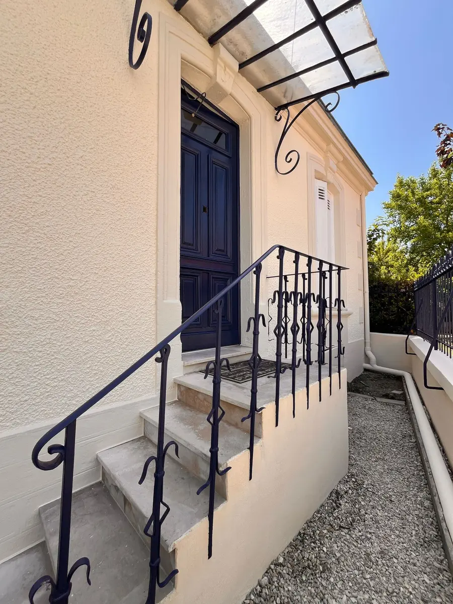Escalier d’entrée avec une rambarde noire et une façade claire.