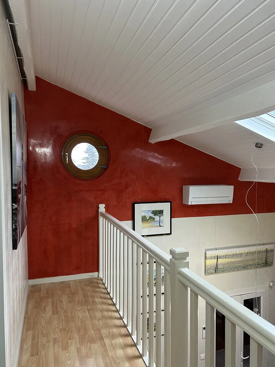 Mur rouge avec une fenêtre ronde au-dessus d’un espace à double hauteur.