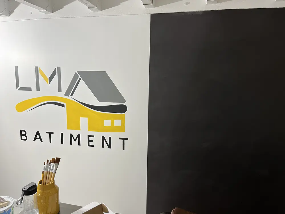 Logo LM Bâtiment peint en gris, jaune et noir sur un mur blanc.