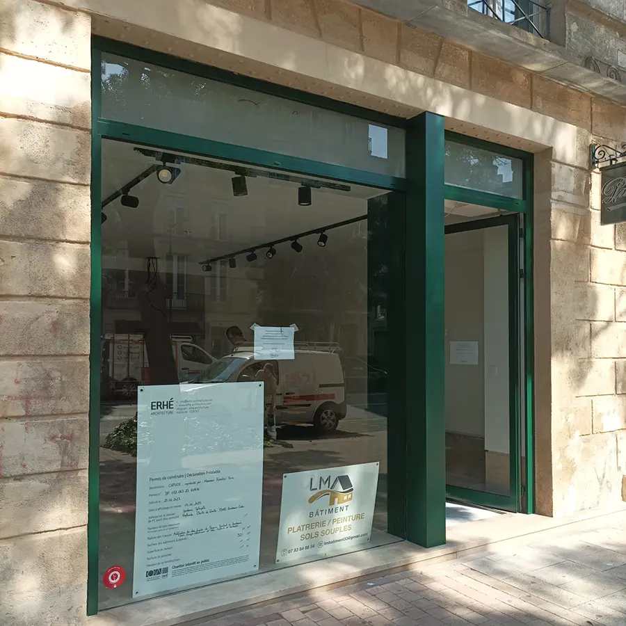 Porte d’entrée vitrée verte d’un commerce avec une affiche LM Bâtiment.
